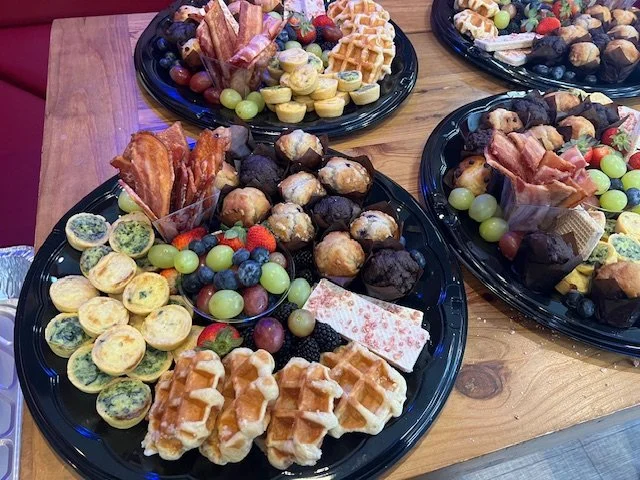 Assorted desserts on black platters, including mini fruit tarts, waffles, chocolate-covered treats, fresh grapes, strawberries, blueberries, and other pastries.