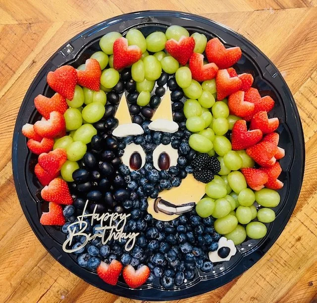 Fruit platter shaped like a butterfly with strawberries, green grapes, black grapes, blueberries, blackberries, and decorative elements, including a "Happy Birthday" message.