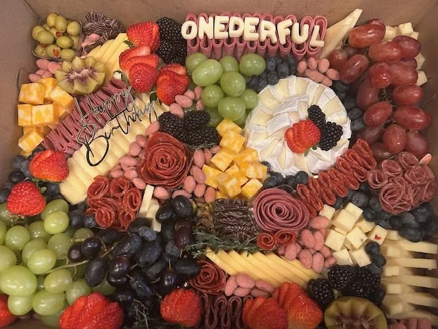 A fruit and cheese platter decorated with various fruits like strawberries, grapes, blackberries, and kiwi, along with cheese slices, cured meats, and garnishes, with decorative pink and white candies spelling 'ONEDERFUL' and 'Sweet Fresh Fruit and C