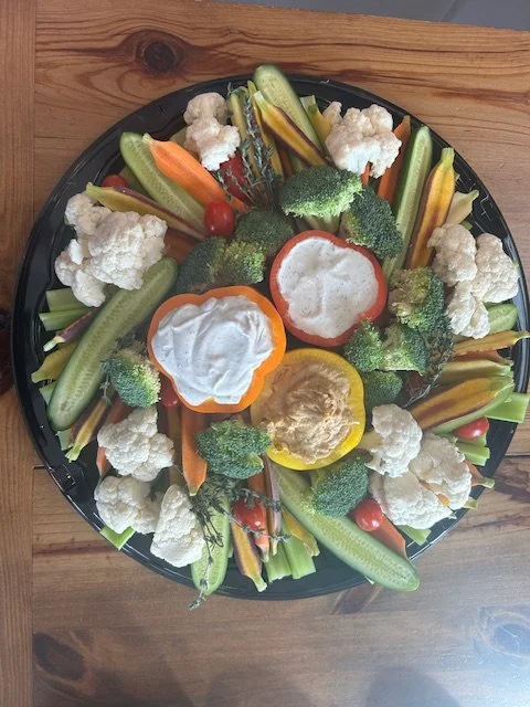 A circular platter of fresh cut vegetables including cauliflower, cherry tomatoes, broccoli, carrot sticks, cucumber slices, and bell pepper rings, placed on a wooden surface.