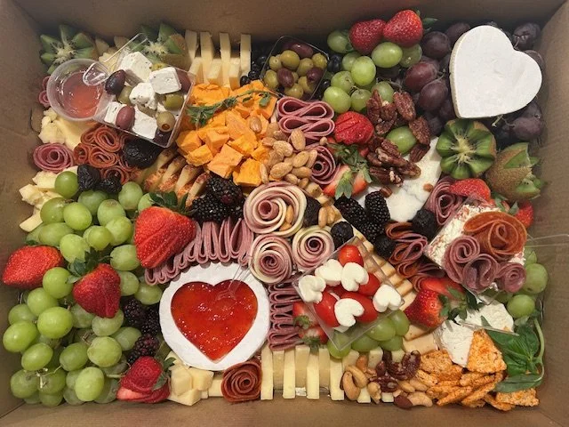 Assorted fresh fruits like strawberries, grapes, and blackberries, along with charcuterie items such as sliced meats, cheese, and nuts, arranged on a tray with decorative elements like a heart-shaped chalkboard and heart-shaped toppings.