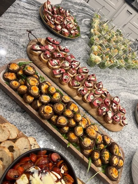 Assorted appetizers including sushi rolls on a wooden serving platter, some topped with sauce and garnishes, alongside a tray of stuffed cherry tomatoes, a bowl of mixed fruit cups with skewers, and a bowl of fruit salad with sliced fruit and dressin