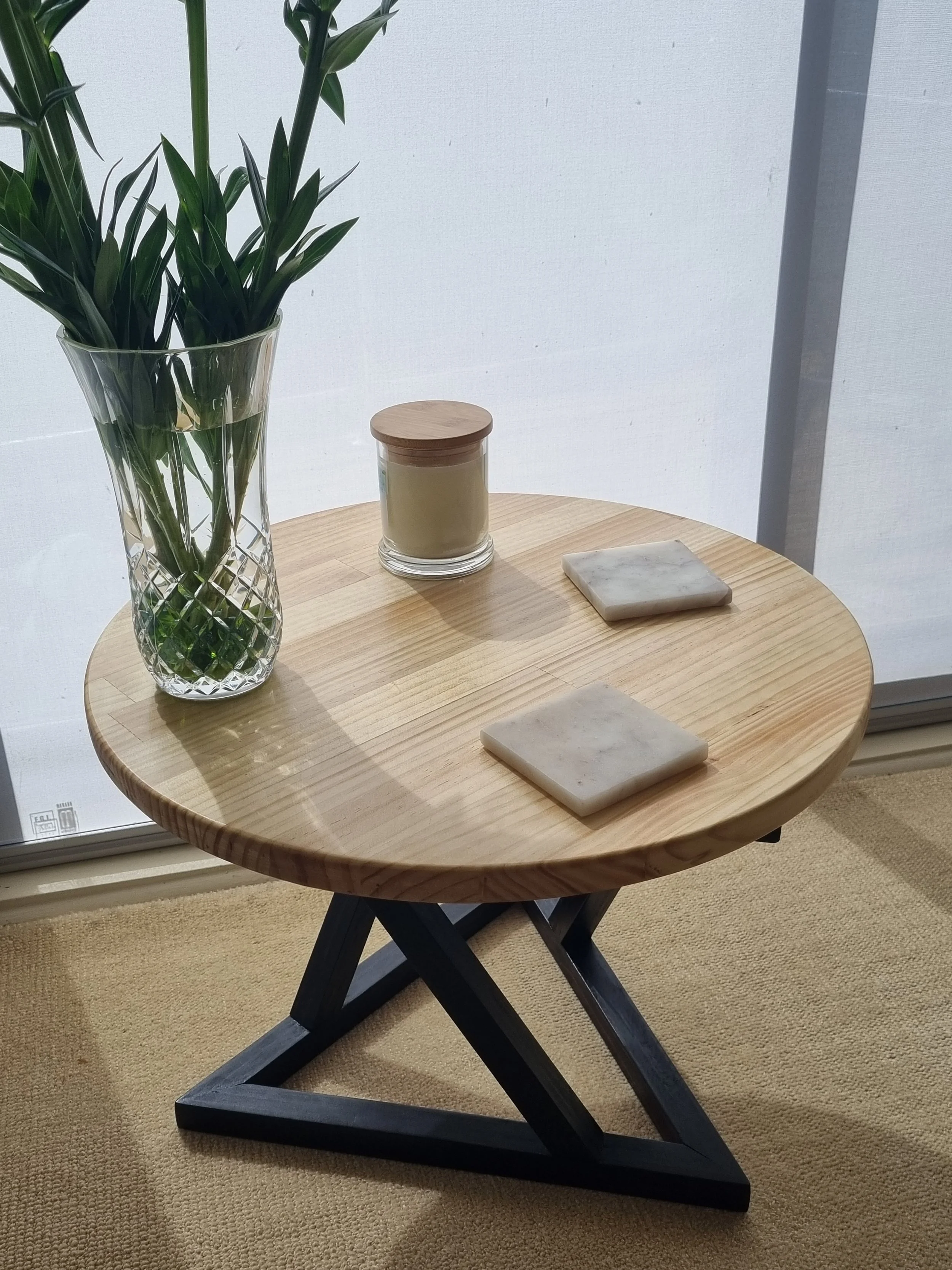 A round wooden table with two marble coasters, a glass vase with green leaves, a candle in a glass jar with a wooden lid, positioned in front of a window with white curtains.