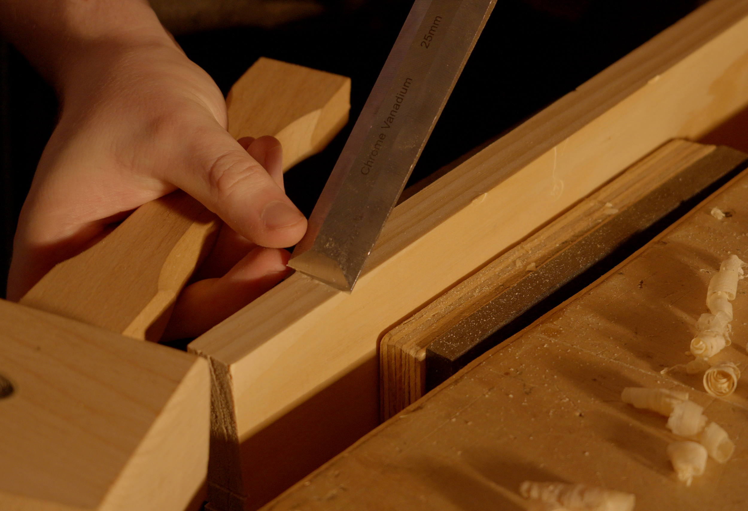 Close-up of a person's hand using a metal chisel to carve or shape a piece of light-colored wood on a workbench, with wood shavings scattered around.