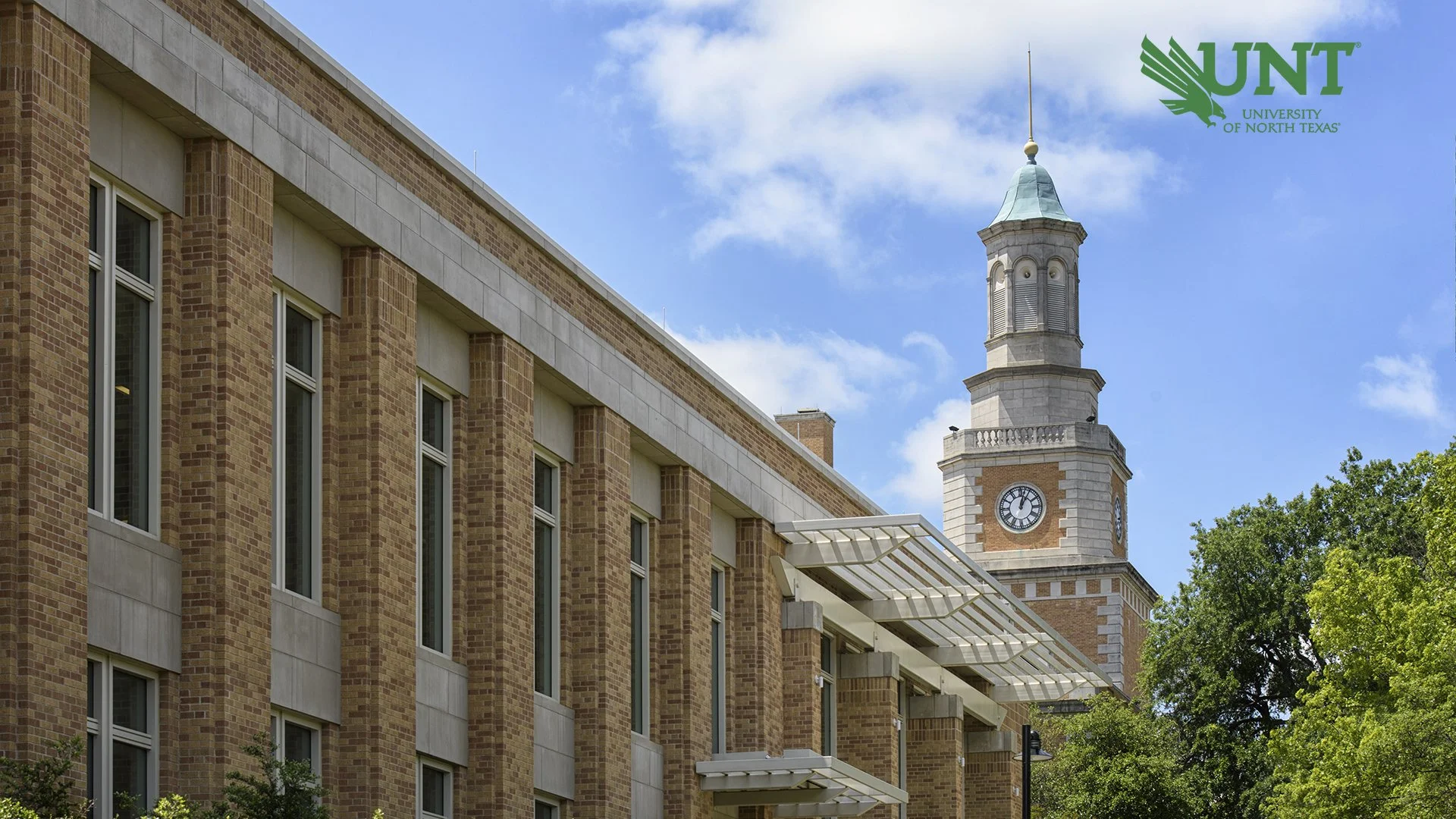 A university building with a brick facade, windows, and an awning. In the background, there is a clock tower with a green roof and a clock face. The sky is blue with some clouds, and there are trees around the building. The UNT logo for the University of North Texas is in the top right corner.