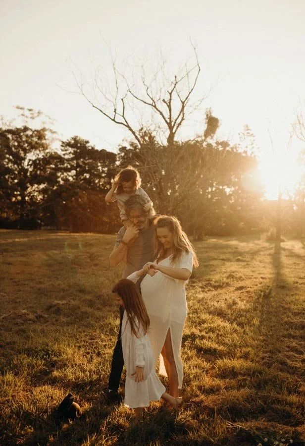 Jade's family of four outdoors at sunset, with a tree in the background, enjoying a beautiful moment. Jade is pregnant and smiling while holding her daughter, who is standing in front. Shael has their son on his shoulders. The scene is warm and illum