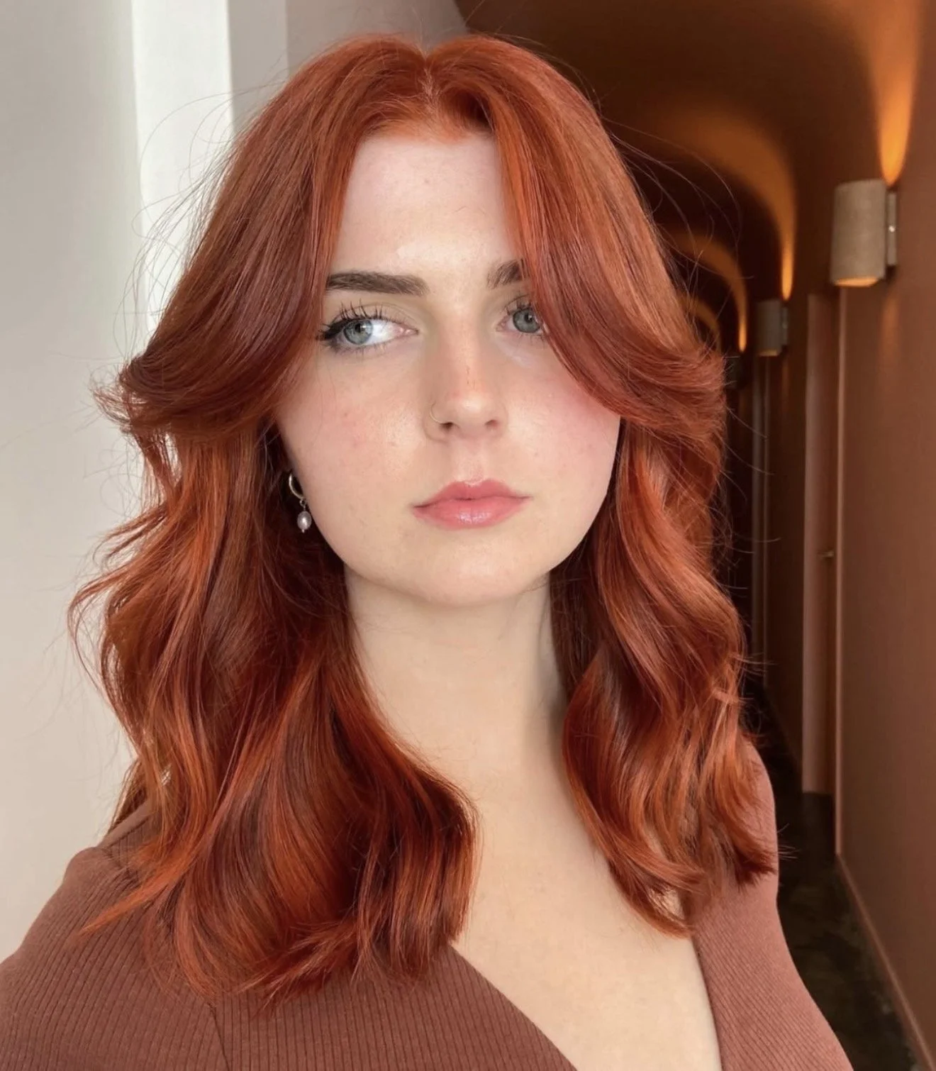 A woman with wavy red hair and light blue eyes poses indoors with warm lighting on the wall behind her.