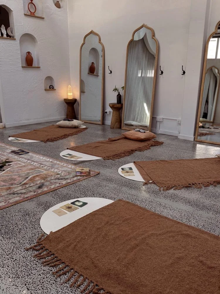 A meditation space with five brown mats and pillows on a speckled floor, three tall ornate mirrors leaning against the wall, small wall shelves with decorative items, and tarot cards on mats.