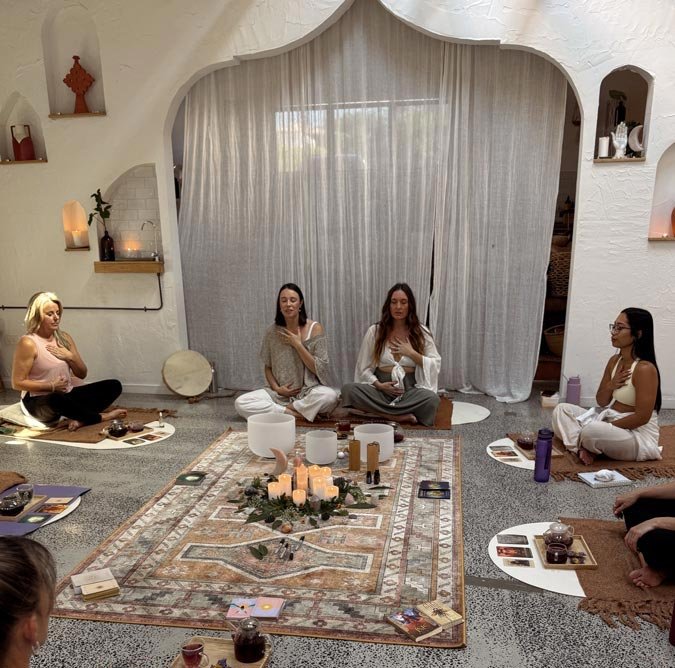 Group of women participating in a meditative, spiritual circle, sitting cross-legged on the floor with candles and tarot cards around a central decorative arrangement.