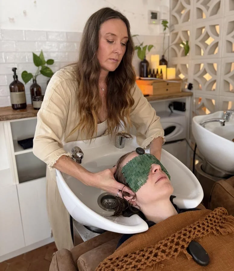 A woman receives a hair treatment at a salon lying back in a wash basin, wearing a green jade eye mask.