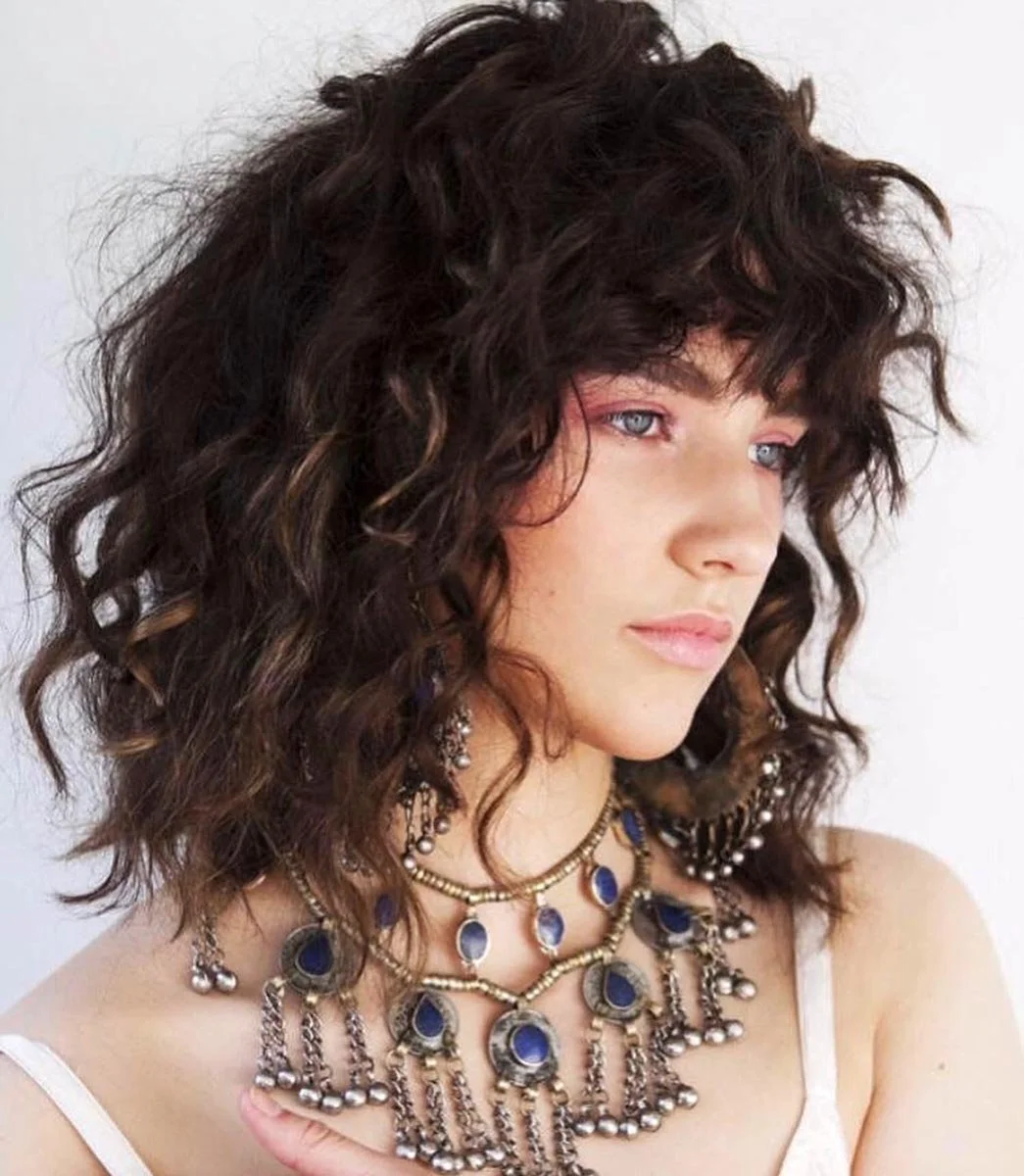 A young woman with curly dark hair and light skin, wearing multiple layered necklaces with blue and silver details, gazing to the side with a neutral expression.