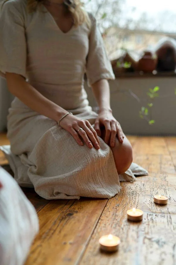 A woman dressed in a light beige dress kneeling on a wooden floor near lit tea light candles, with a window and houseplants in the background.