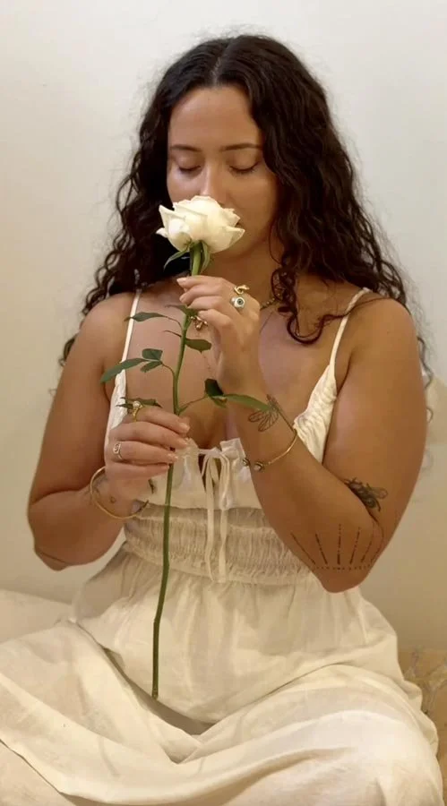 Woman with dark curly hair holding a white rose close to her face with eyes closed, wearing a white dress, sitting against a plain wall.