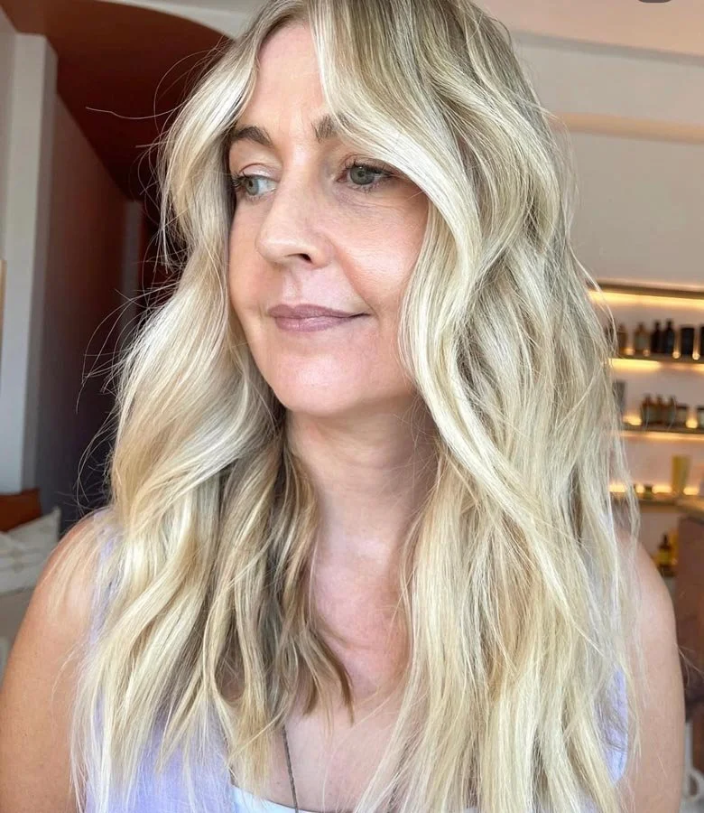 A woman with long, wavy blonde hair looking to her left in a modern room with shelves and warm lighting in the background.