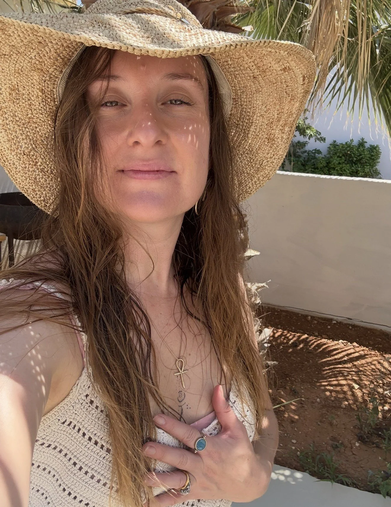 Jade wearing a large straw hat, necklace with an ankh pendant, standing outdoors in bright sunlight, with palm trees and a white wall in the background.