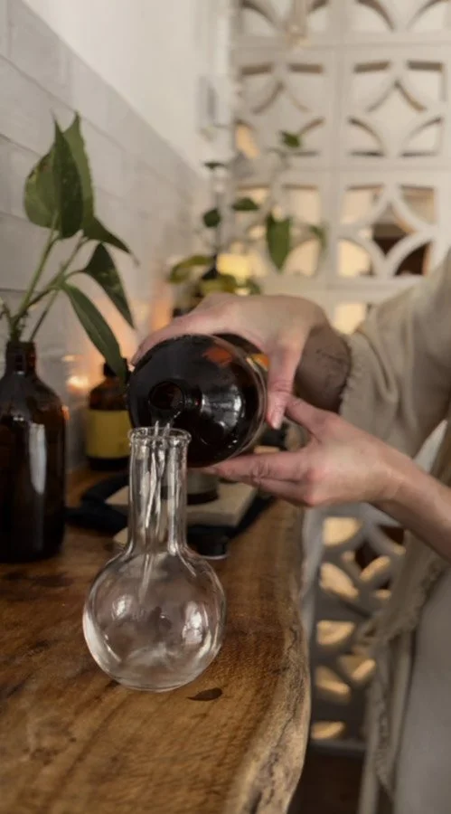 Person pouring a clear liquid into a glass flask on a wooden table with indoor plants and decorative bottles in the background.
