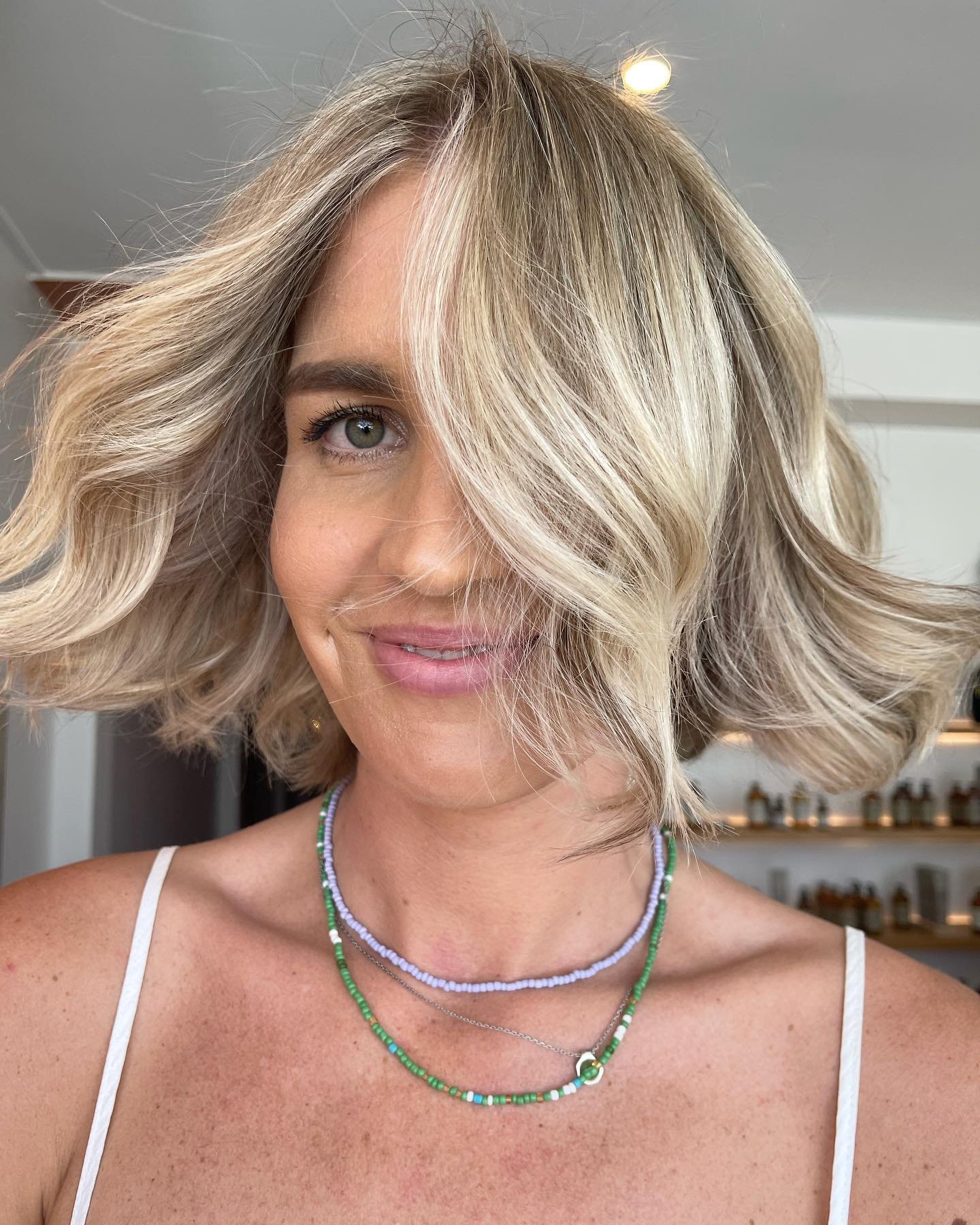 A woman with blonde, wavy hair styled in a chin-length bob, wearing a white spaghetti strap top and layered beaded necklaces, smiling softly.