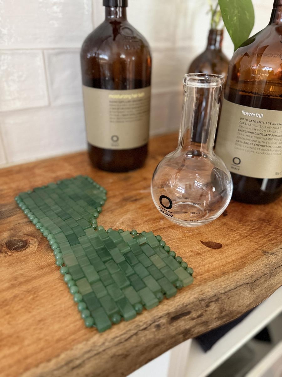 Decorative green jade eye mask, a clear glass flask, and three amber bottles with labels are arranged on a wooden surface, with a white brick wall in the background.