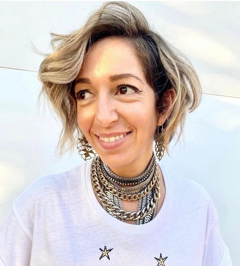 A woman with short, wavy blonde and dark hair smiling at the camera, wearing a white T-shirt with star graphics and multiple layered gold and silver necklaces along with earrings, against a plain light background.