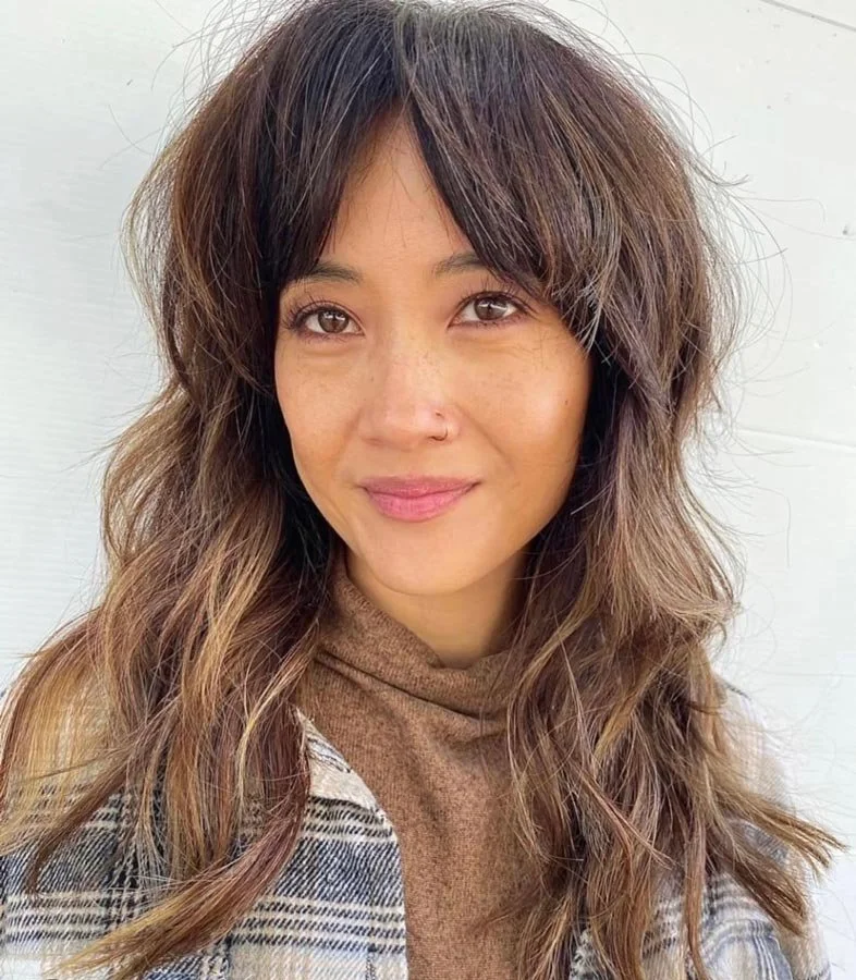 A woman with wavy brown hair, light brown eyes, and a nose piercing, smiling slightly, wearing a brown sweater and a plaid coat, against a plain light background.