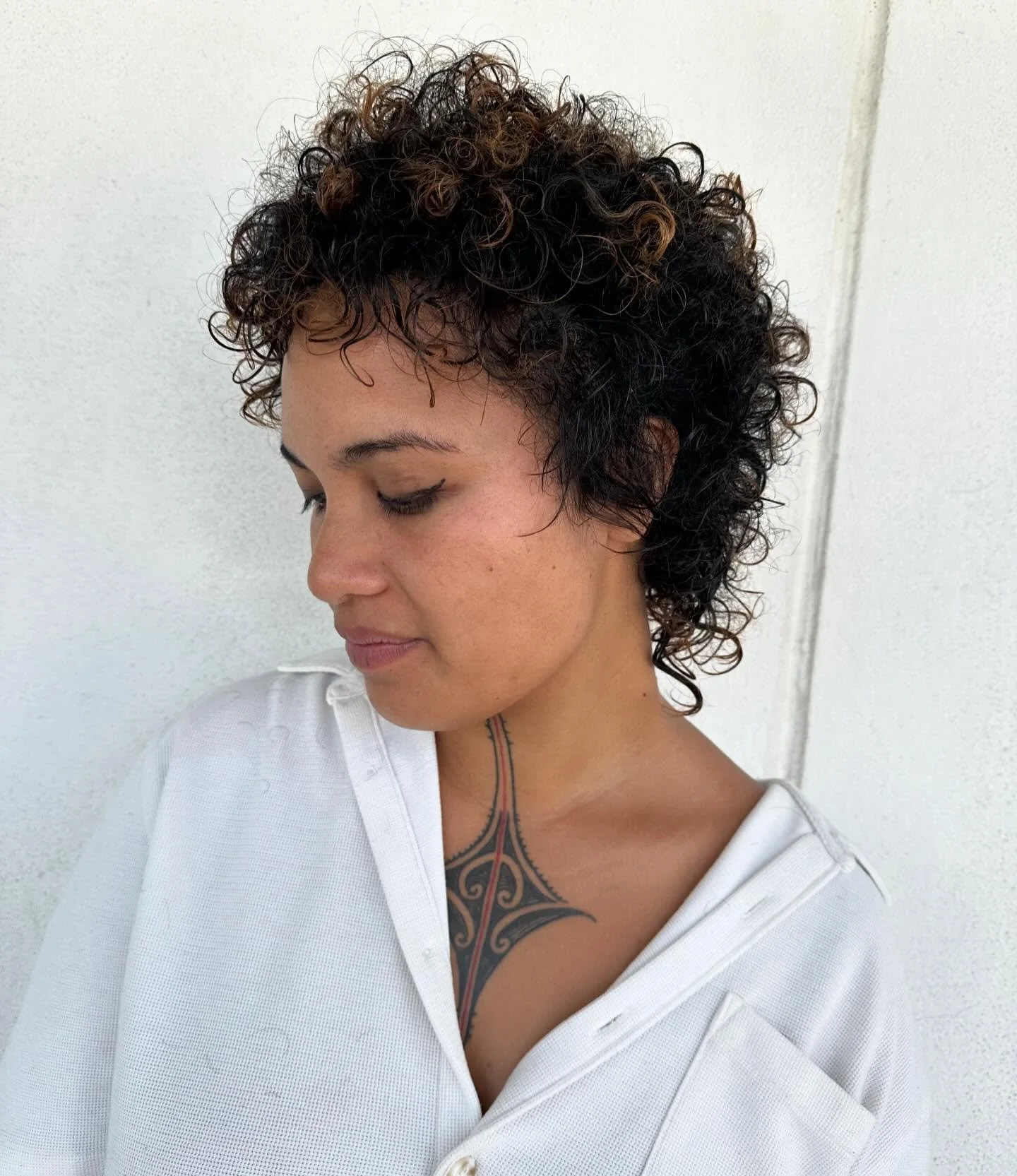 A woman with short, curly dark hair with lighter tips, looking down, wearing a white shirt and a visible tribal tattoo on her chest.
