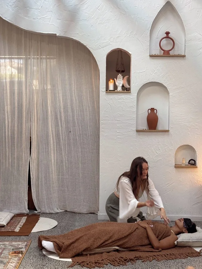 A woman lying on a mat receiving an energy, healing therapy from Jade in a cozy room with white textured walls and decorative shelves with vases and candles.