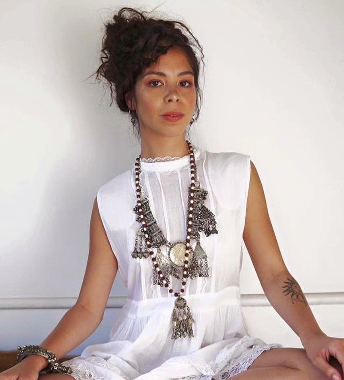 A woman with curly hair in an updo, wearing a white sleeveless dress and layered bohemian jewelry, sitting against a plain white wall.