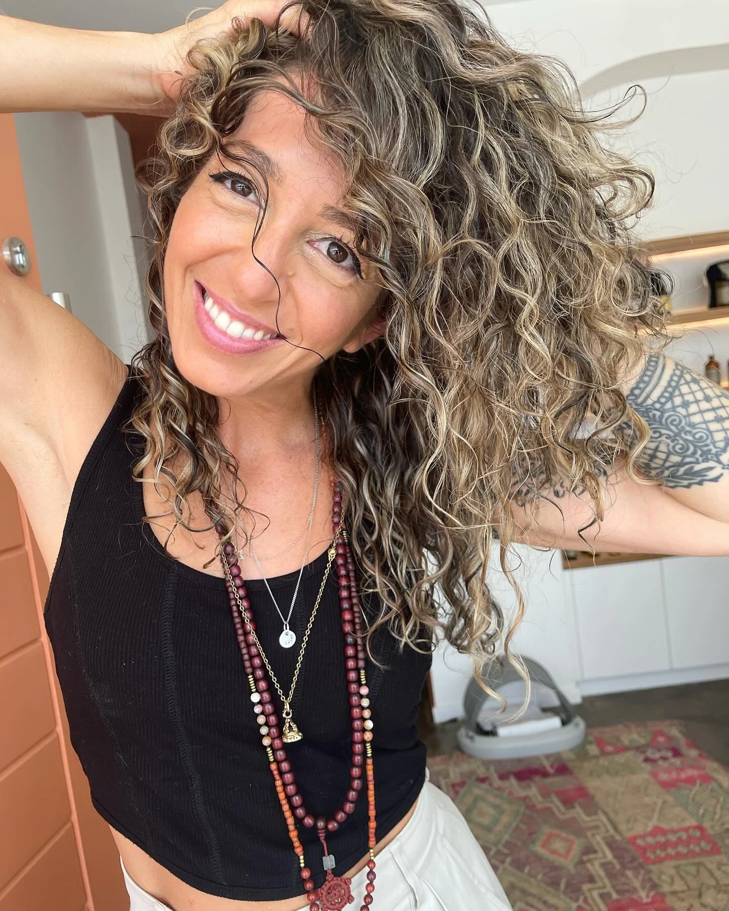 A woman with curly hair smiling, wearing a black sleeveless top and multiple layered necklaces, standing in a room with shelves and a patterned rug.