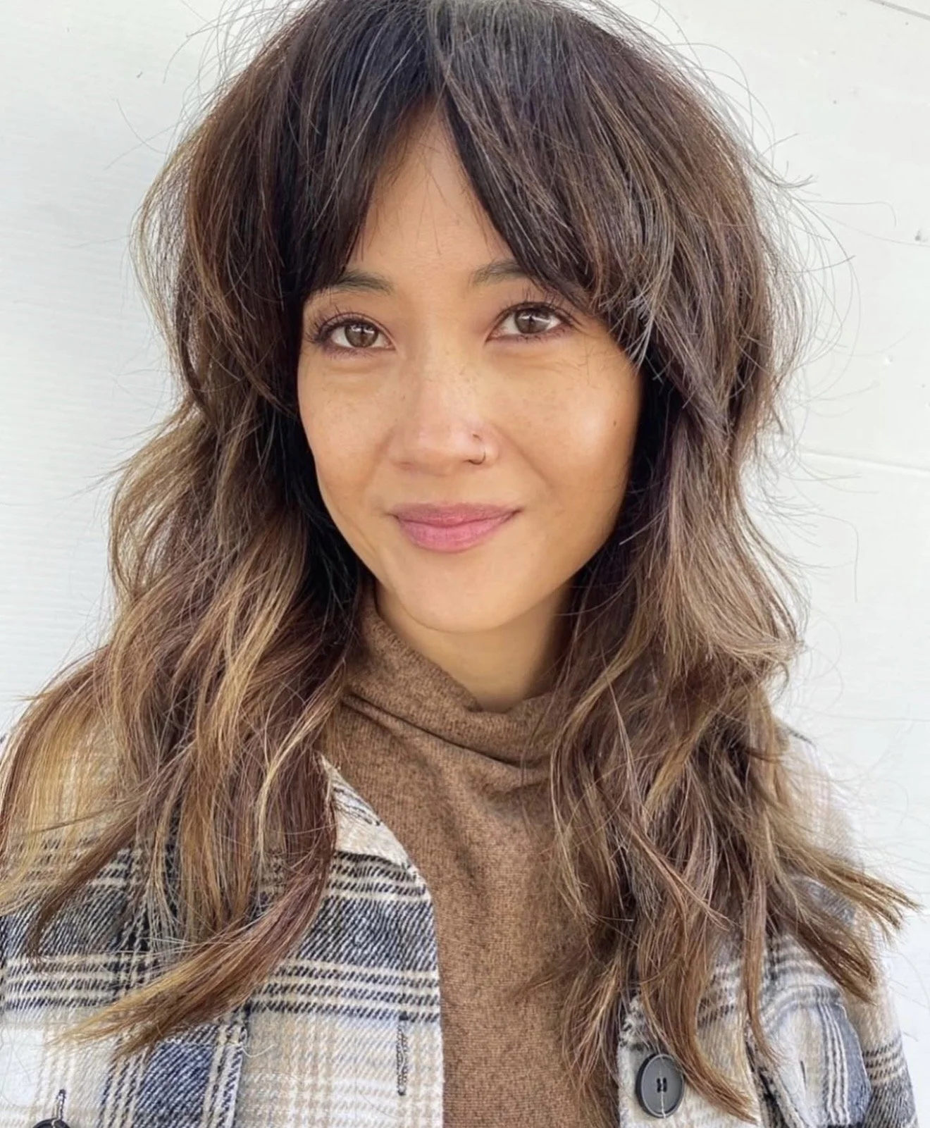 A woman with wavy brown hair and light skin smiling, wearing a brown turtleneck and a plaid jacket, standing against a white wall.