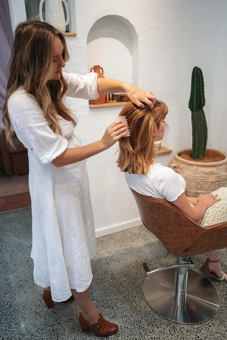 A woman is getting her hair styled by Jade in the salon. Jade is standing and combing the woman's hair with a rose quartz comb, which is shoulder-length and reddish-brown. The woman is seated in a brown salon chair, wearing a white shirt. The salon