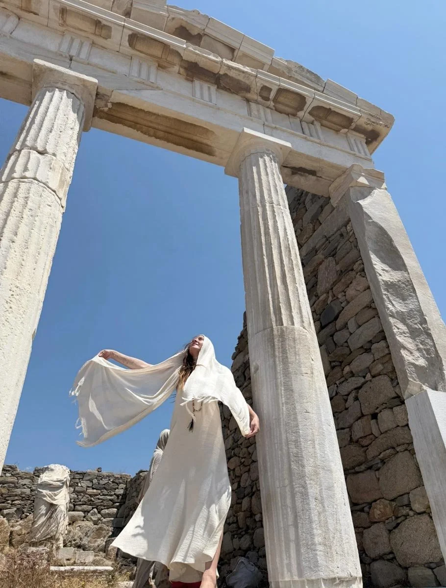 Jade in a white dress and matching headscarf standing underneath ancient Greek columns at a historic site, with clear blue sky in the background.