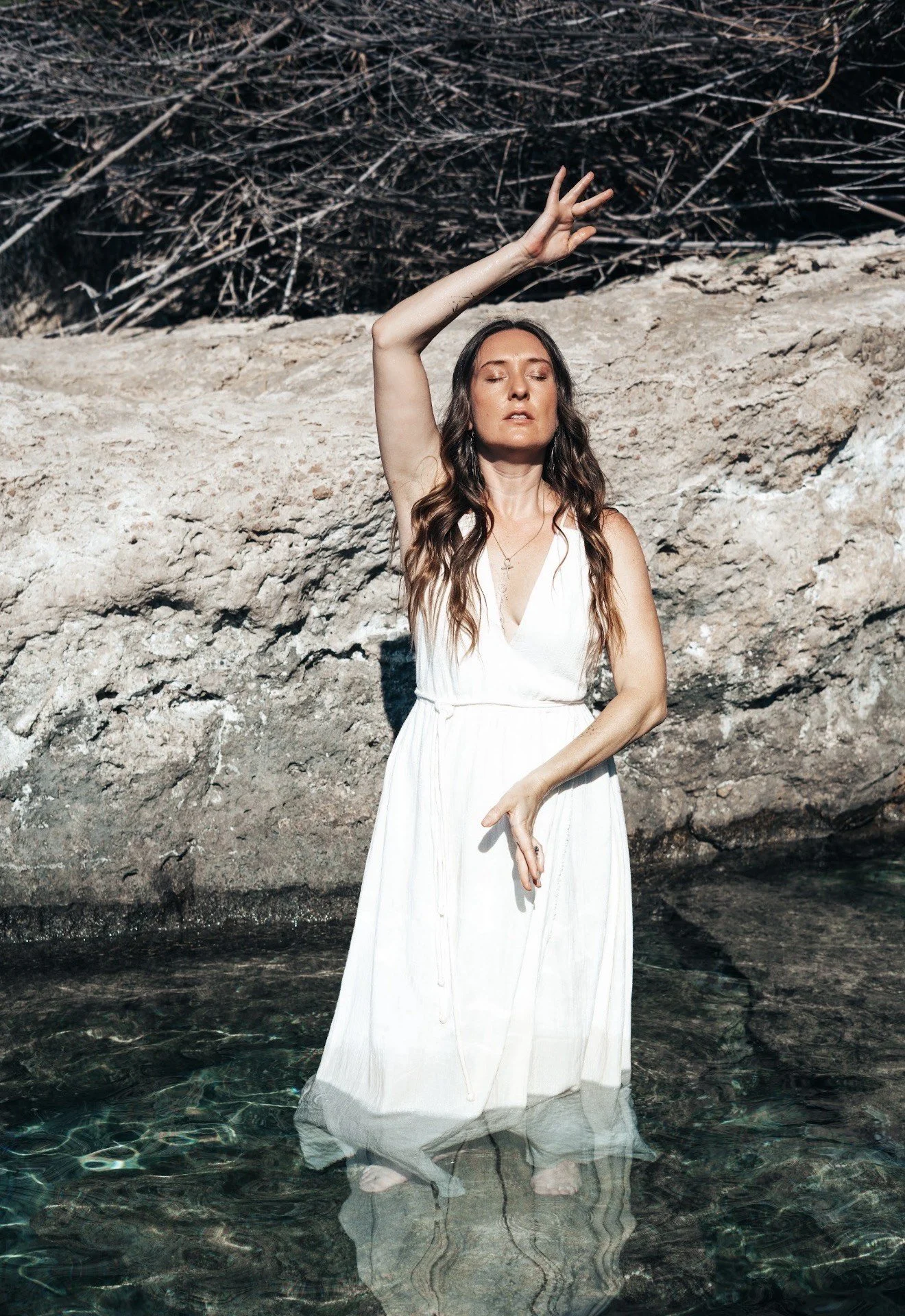 Jade in a white dress standing in shallow water with her eyes closed, one arm raised above her head, and the other hand near her waist, against a rocky background.