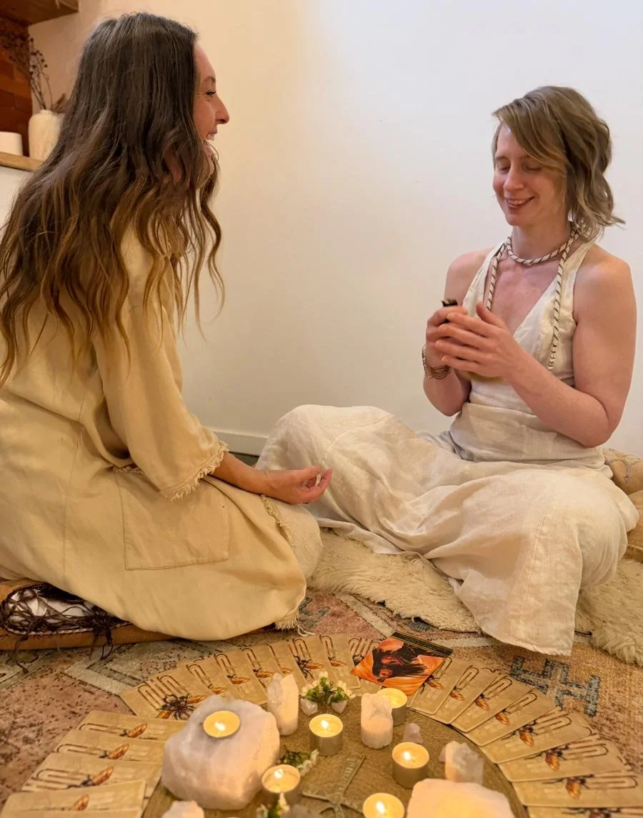 Jade sits on the floor facing another woman, engaging surrounded by lit candles and crystals.