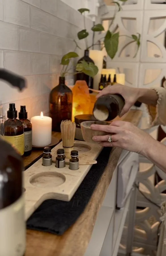 Someone pouring a powdered substance into a small bowl on a wooden spa tray, with essential oils, candles, and plants on a countertop in the background.