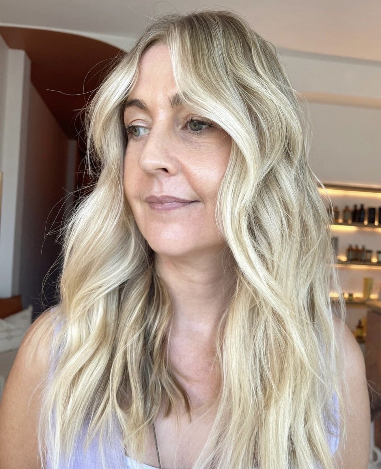 Woman with long blonde wavy hair and natural makeup, indoors with shelves in the background.