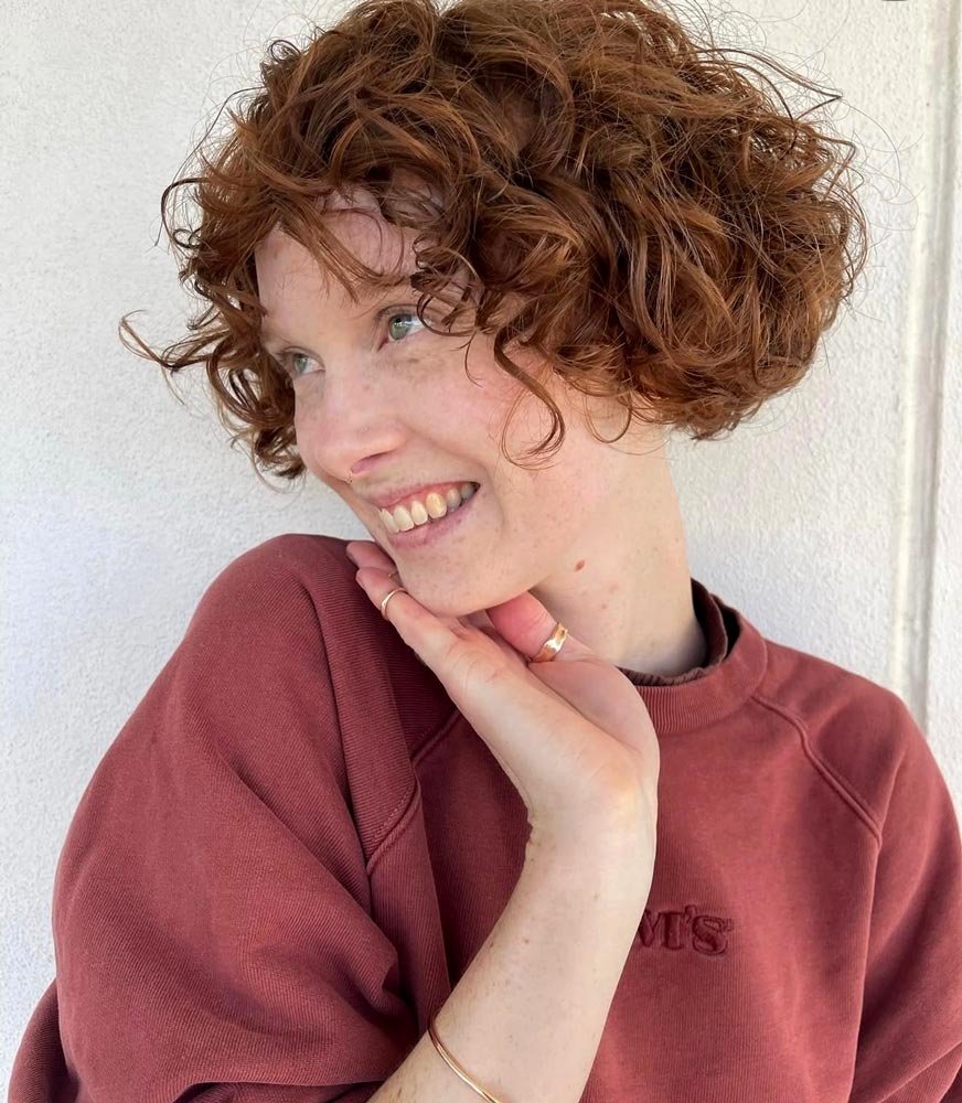 A young woman with curly red hair smiling and looking to the side, wearing a maroon sweatshirt, standing against a white wall.