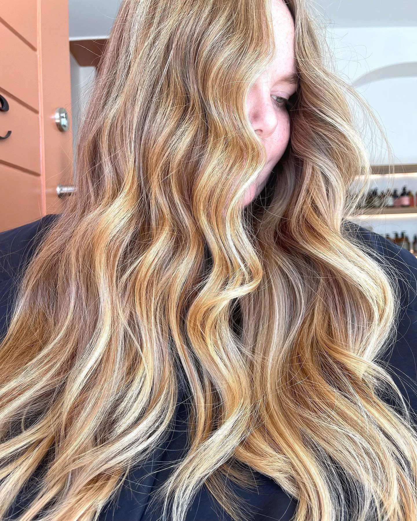 Close-up of a woman with long, wavy, blonde hair with darker roots, looking down with her face partially covered by her hair.