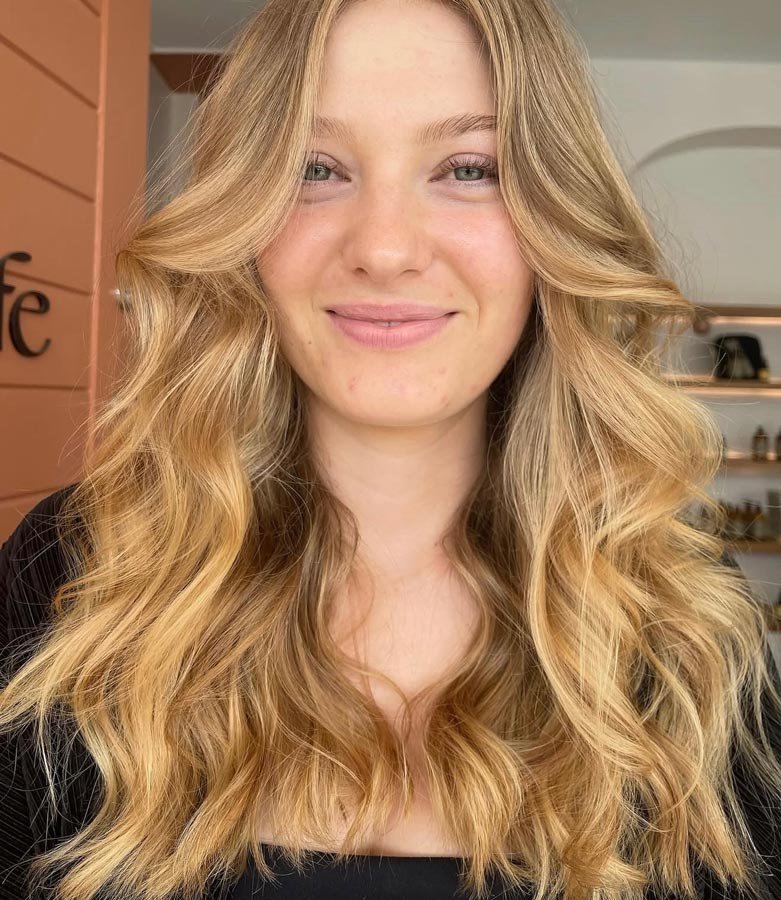 A young woman with long, wavy blonde hair smiling at the camera, in a natural indoor setting.