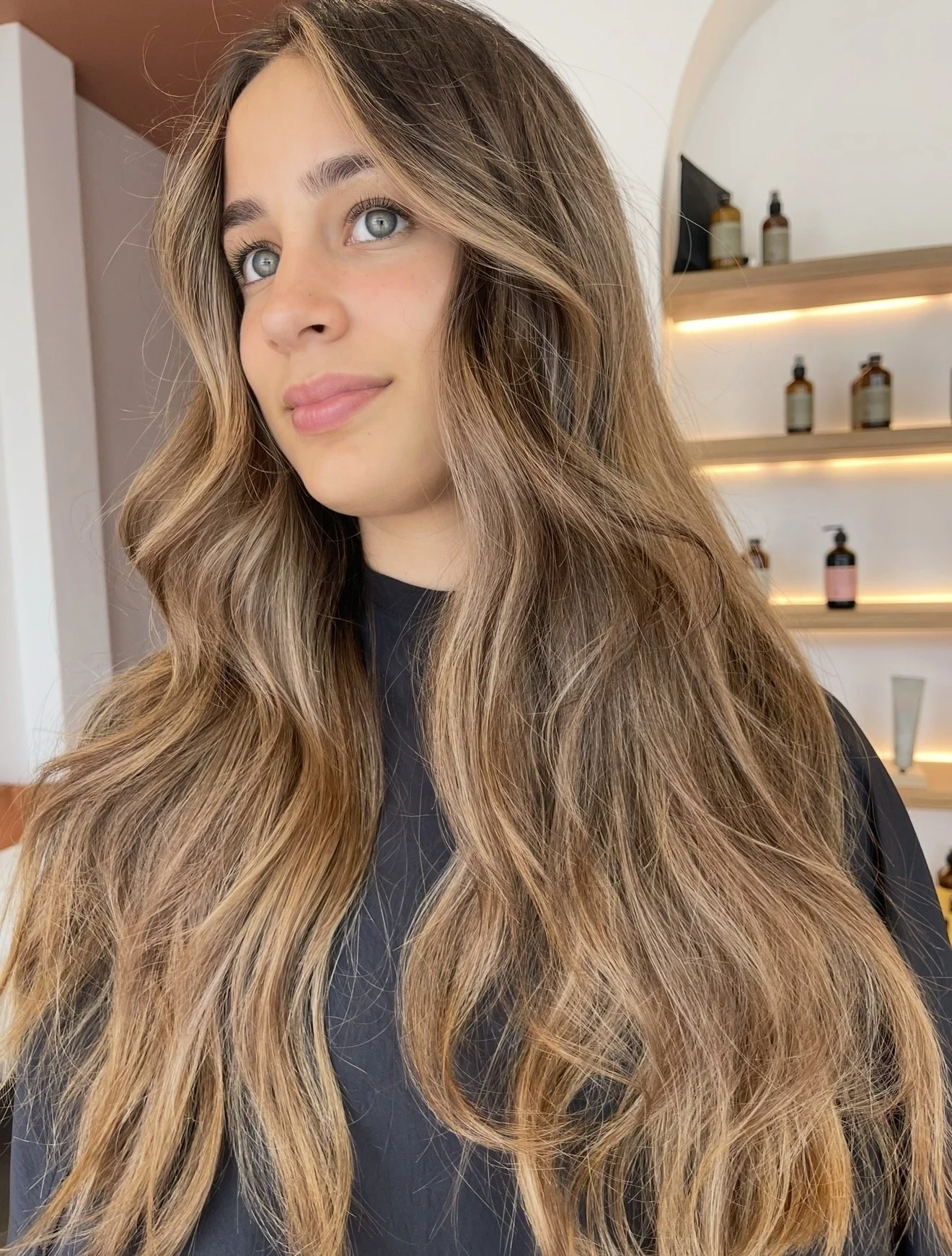 A woman with long, wavy, light brown hair with blonde highlights, wearing a black top, standing indoors in front of a wall with shelves of small bottles and containers.