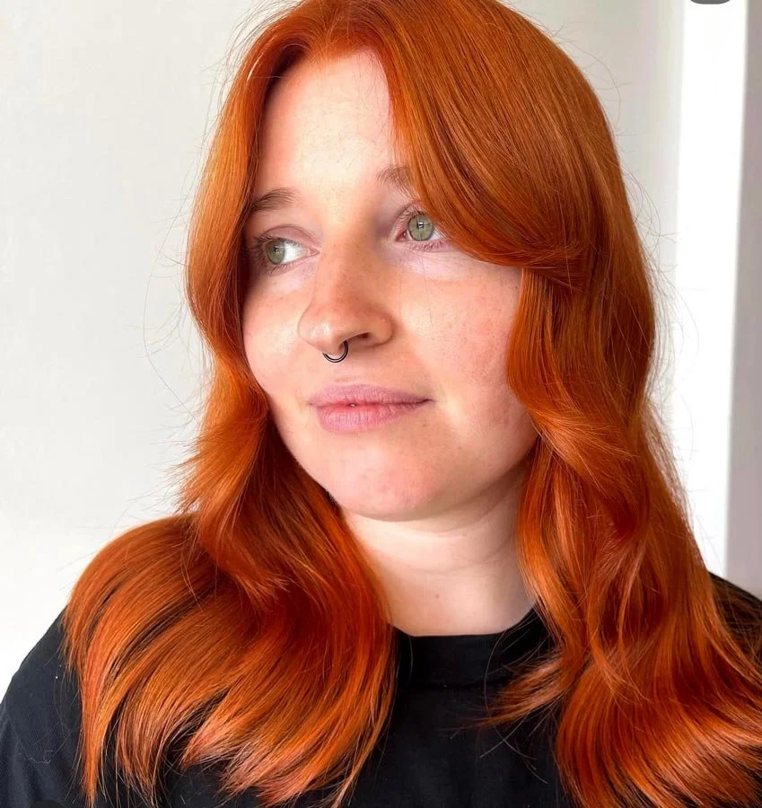 A young woman with long, wavy red hair and green eyes, wearing a black top and a septum ring, looking to the side against a plain light-colored background.