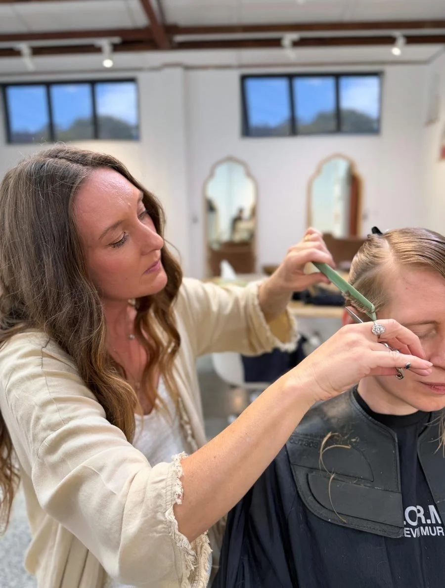 Jade giving a haircut to a woman in a salon with mirrors and windows in the background.