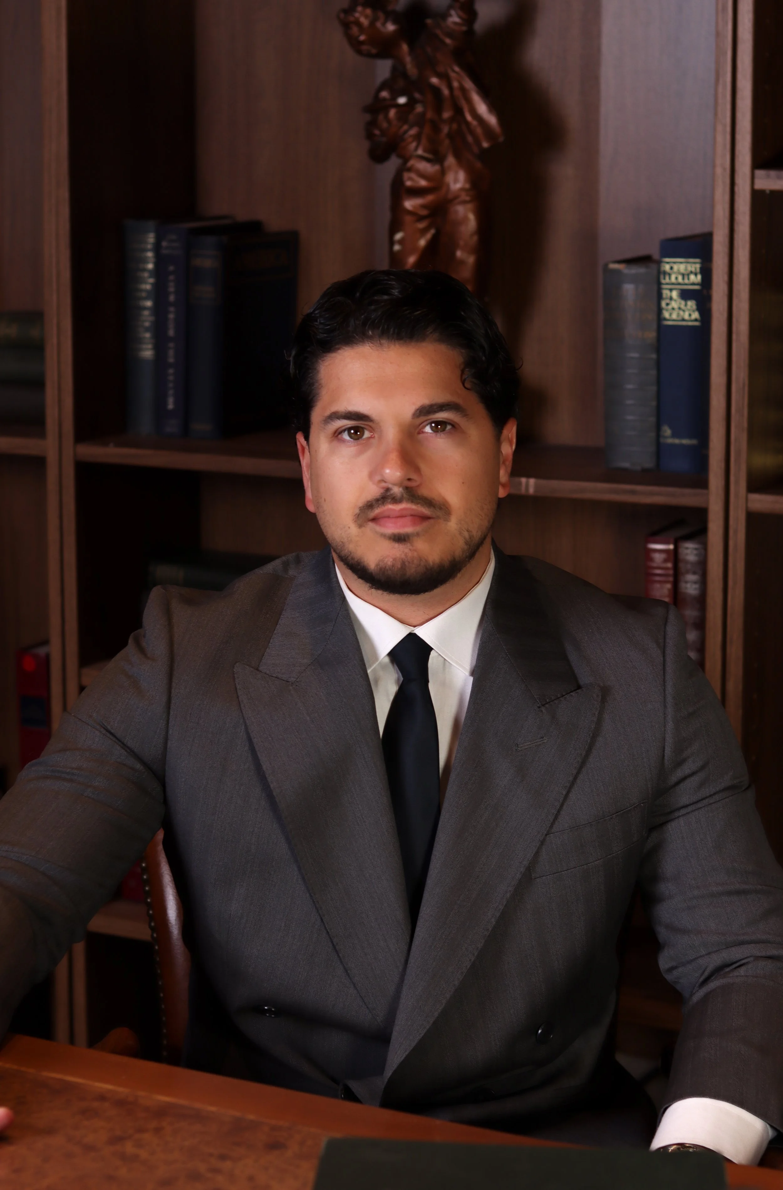 A man in a gray suit sitting at a wooden desk in front of a bookshelf with books and a wooden sculpture of a person.