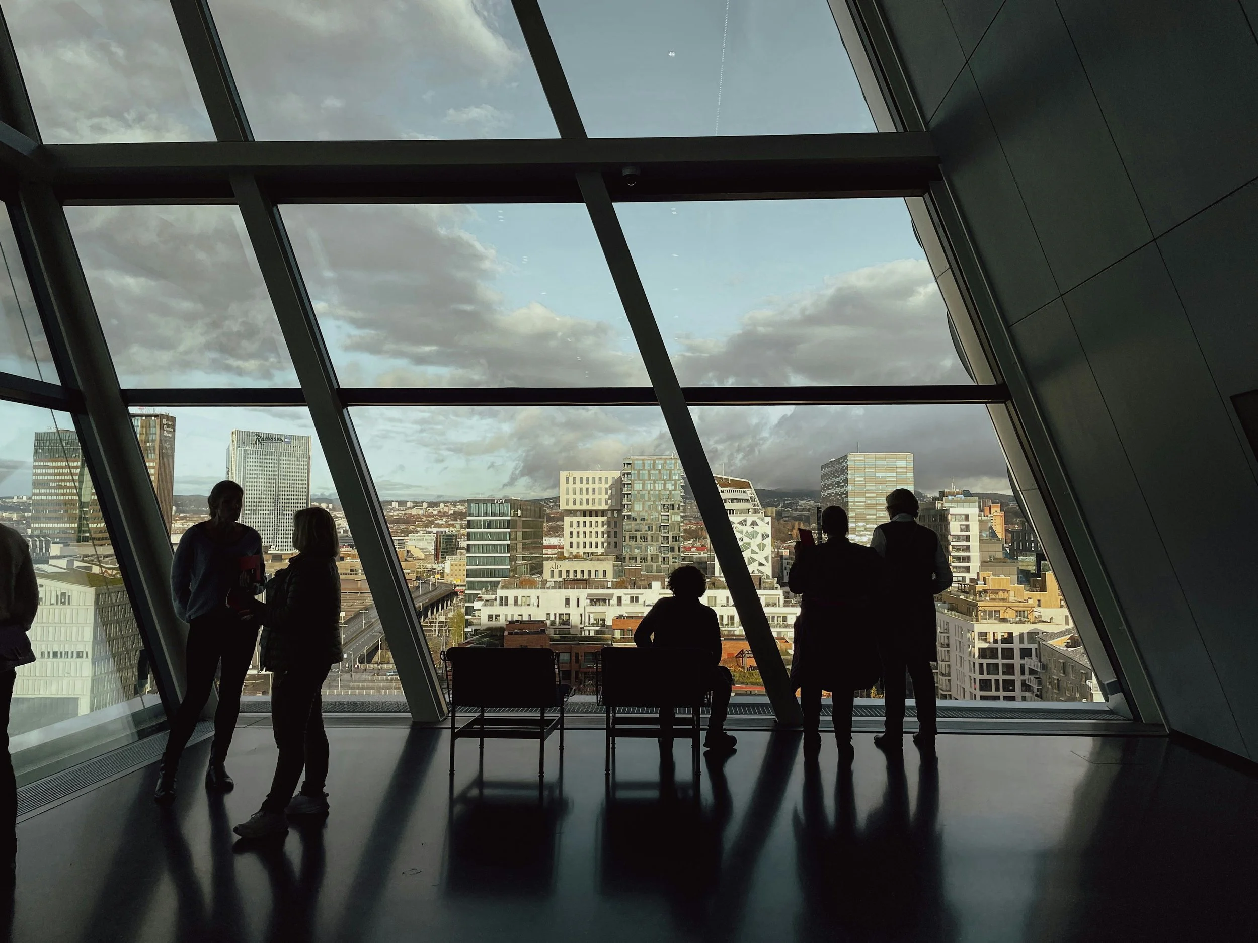 Silhouettes of people inside a modern building with large angled glass windows, overlooking a city skyline with tall buildings and cloudy sky outside.