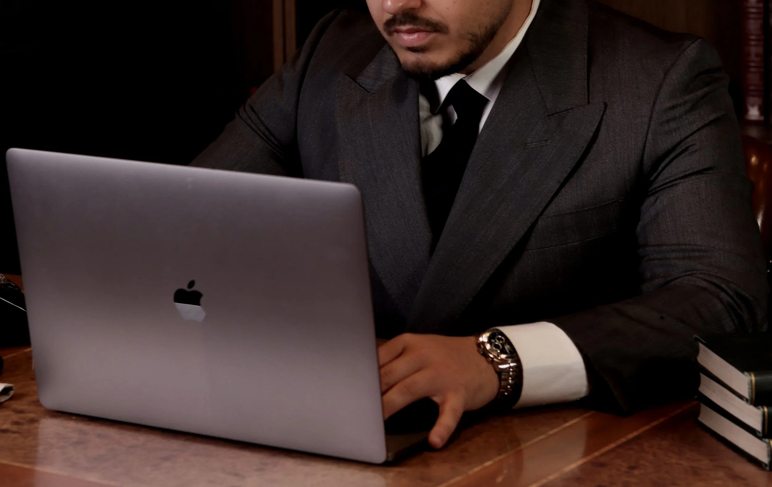 A man in a dark suit working on a silver Apple laptop, sitting at a wooden desk with a few books and a phone nearby.