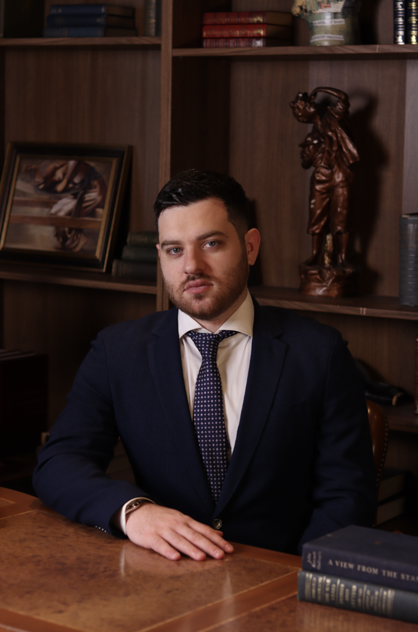 A man in a dark suit and tie sitting at a wooden desk in a library or study, with wooden bookshelves behind him containing books and a wooden sculpture.