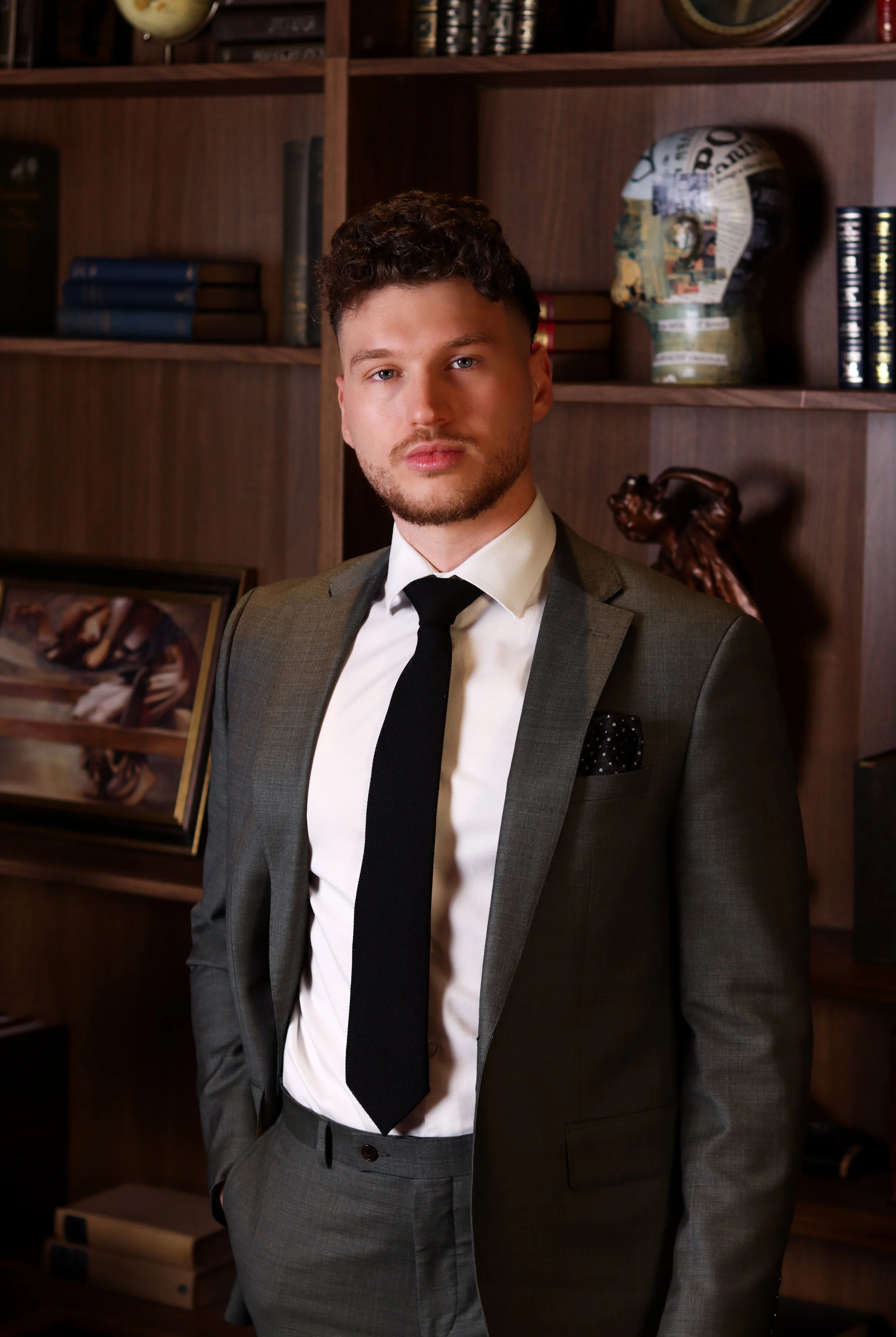 A young man in a gray suit and tie standing in front of a wooden bookshelf with decorative items and framed pictures.