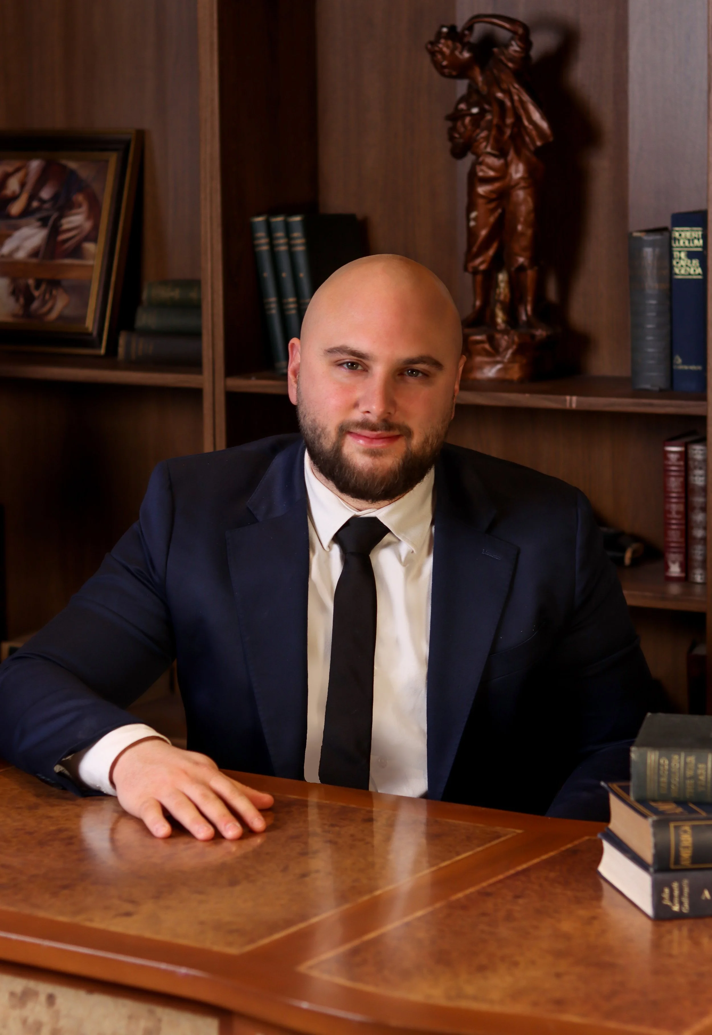 A man in a navy suit, white shirt, and black tie sitting at a wooden desk with books on it, in front of a wooden bookshelf with a carved sculpture and framed photo in the background.