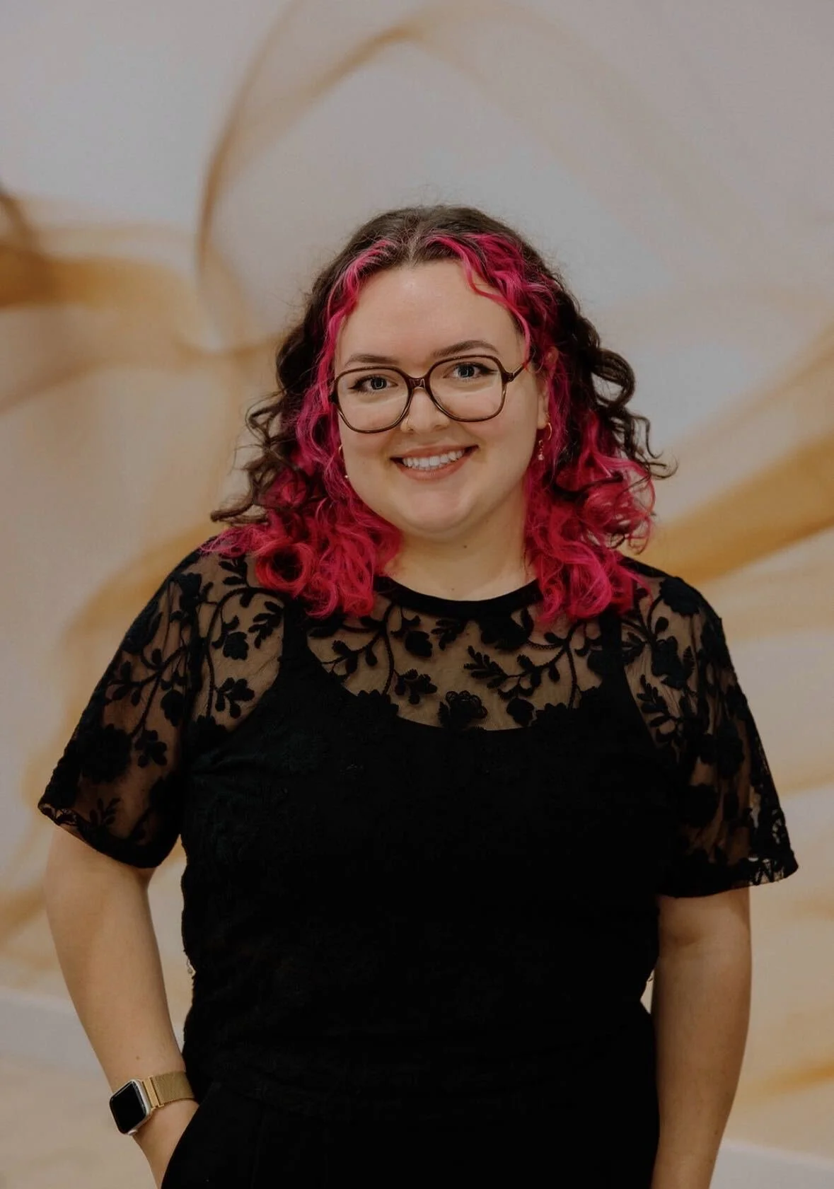 A woman with long, wavy brown hair, wearing glasses and a tan short-sleeve top, smiling at the camera against a light-colored textured wall background.