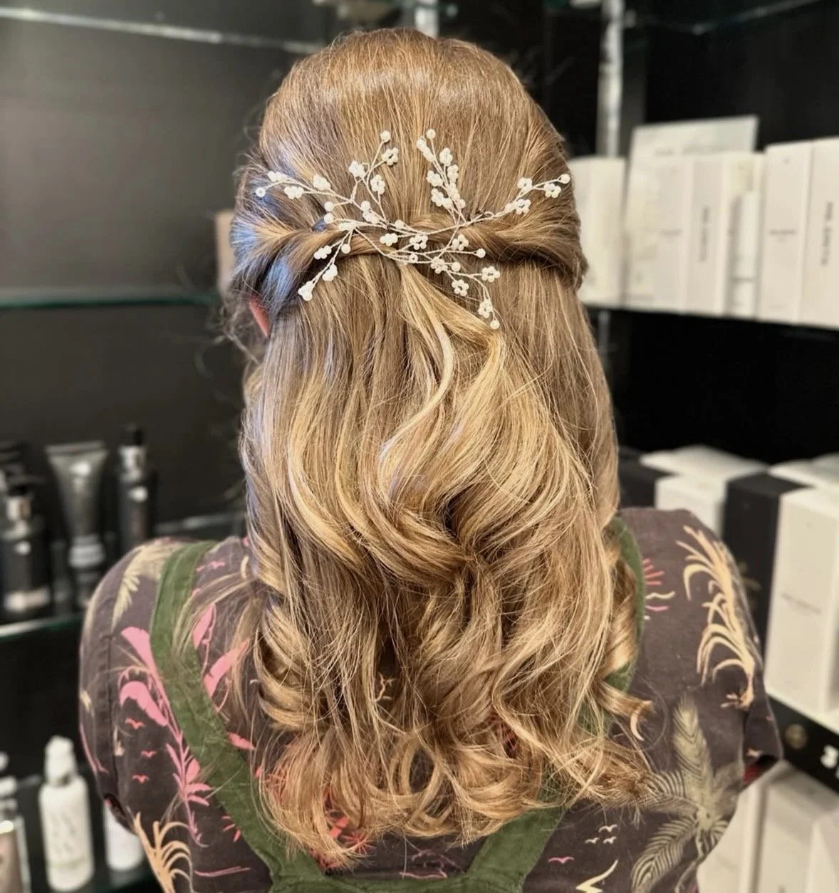 Back view of a woman with long, wavy blonde hair, adorned with a white floral hairpiece, wearing a dark shirt with a colorful floral pattern, in a store setting with shelves of beauty products.
