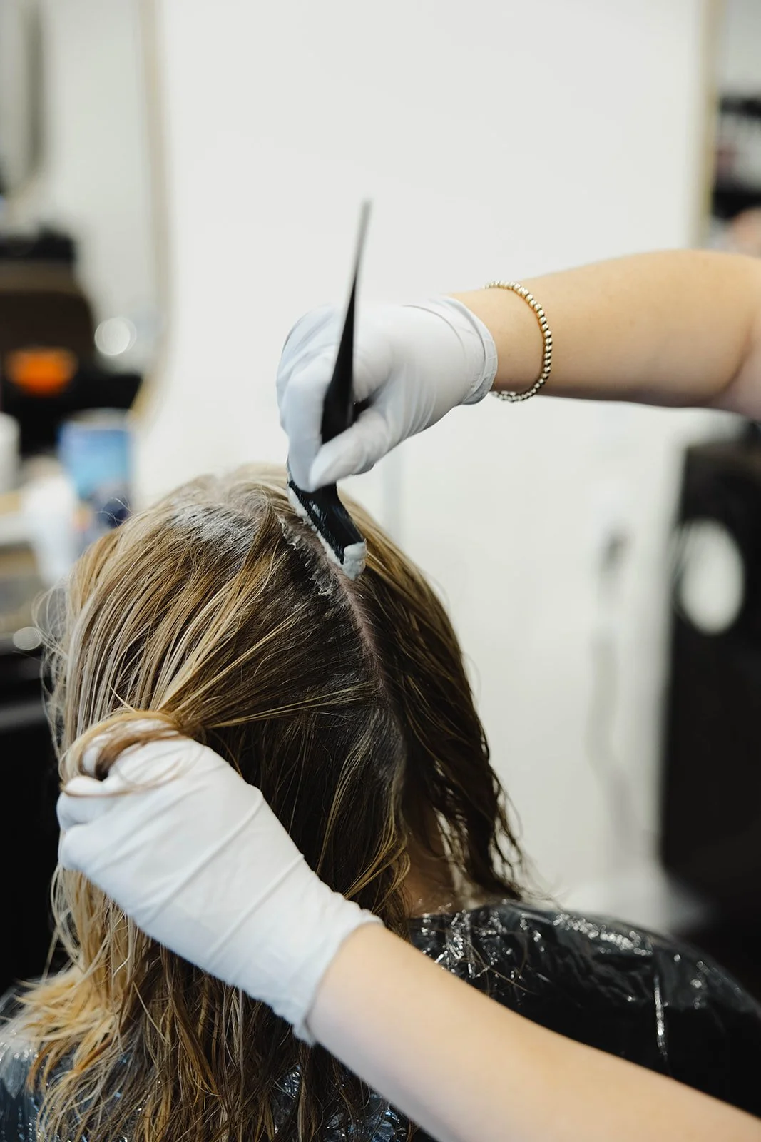 A hairdresser applies hair color to a woman's hair in a salon, wearing white gloves and a bracelet.